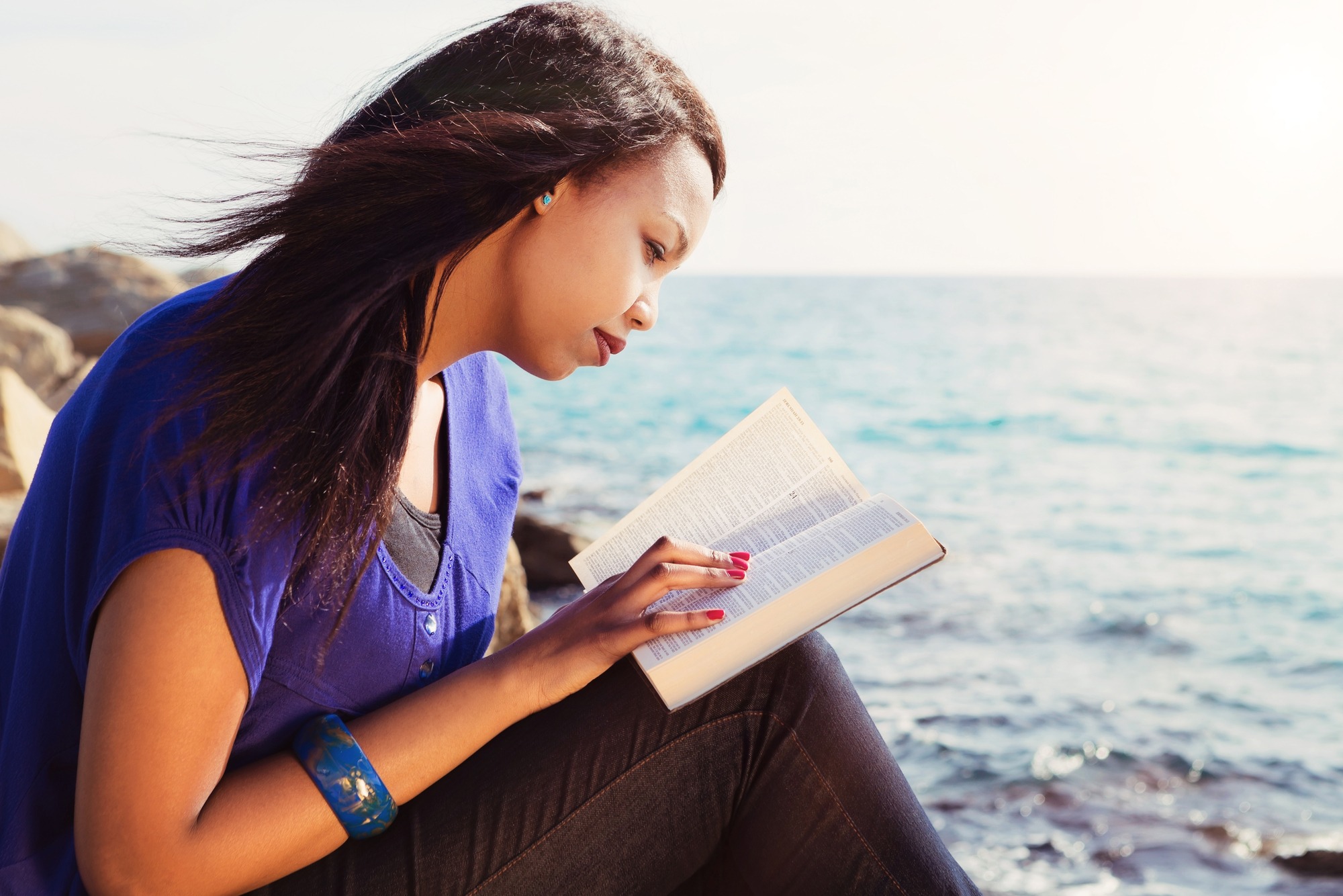 A woman reads a book by the water, with sunlit waves in the background, capturing a serene moment of reflection and relaxation.