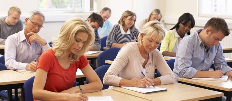 Adults participating in an examination, focused on their papers in a classroom setting. The diverse group illustrates a learning environment.