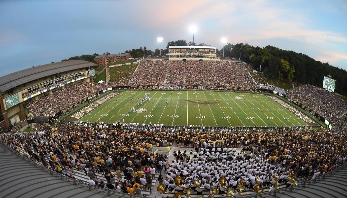Packed stadium during a nighttime football game, with fans cheering as players compete on the field under bright floodlights.