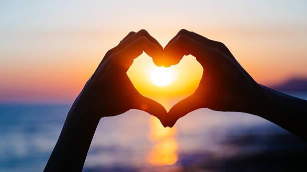 Hands forming a heart shape against a sunset backdrop, symbolizing love and warmth.